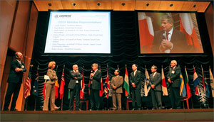 Eight people standing on a stage with flags from various countries in the background.